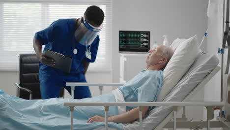 An-elderly-male-patient-lying-in-a-hospital-bed-connected-to-an-ECG-machine-talks-to-a-black-doctor-in-a-protective-mask
